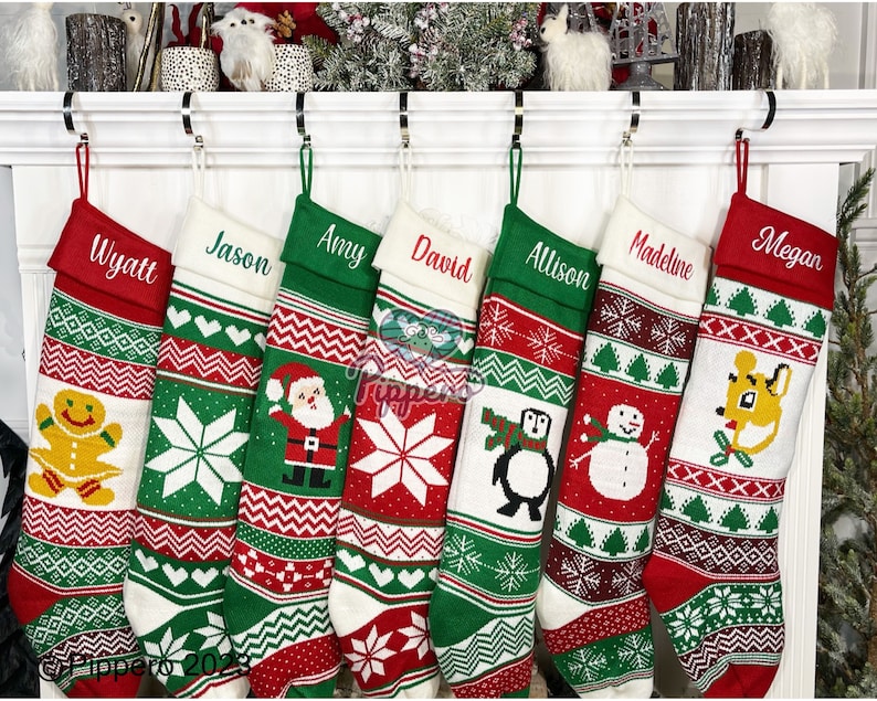 May include: Seven personalized Christmas stockings with red, green, and white knitted patterns. The stockings feature various holiday designs, including a gingerbread man, a snowflake, Santa Claus, a penguin, a snowman, a reindeer, and a deer. The stockings are hung on a white fireplace mantel.