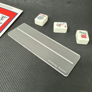 May include: A rectangular, clear acrylic Mahjong tile rack featuring a central horizontal groove and the text Mahjong Master etched in the bottom right corner, accompanied by three white Mahjong tiles with colorful symbols and the word Joker.
