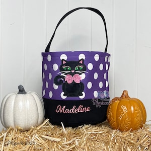 May include: Purple and white polka dot trick or treat bag with a black cat with pink bow and green eyes. The bag has a black bottom with the name Madeline embroidered in pink.
