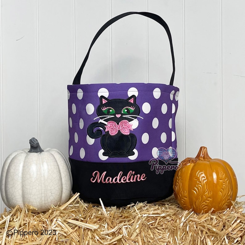 May include: Purple and white polka dot trick or treat bag with a black cat with pink bow and green eyes. The bag has a black bottom with the name Madeline embroidered in pink.