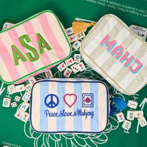 Könnte beinhalten: Drei gestreifte Mahjong-Spieltaschen in Rosa, Beige und Blau, jeweils mit gestickten Details. Eine Tasche hat die Initialen "ASA", eine andere "MAHJ", und die dritte zeigt ein Friedenszeichen, ein Herz und ein Blumenmuster mit dem Text "Peace, Love, & Mahjong".