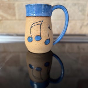 May include: A handmade ceramic mug with a blue rim and handle. The beige body features black musical notes with blue accents. The mug is designed for hot beverages.