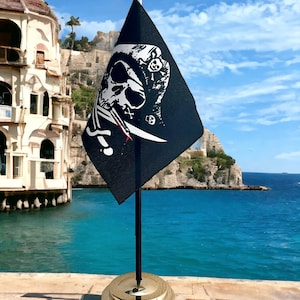 May include: A black pirate flag with a white skull and crossbones design. The flag is on a gold metal stand with a black pole. The flag is in front of a blue body of water.