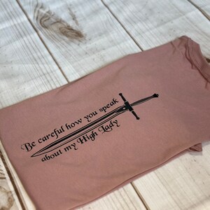 May include: A mauve t-shirt with a black graphic of a sword and the text "Be careful how you speak about my High Lady".