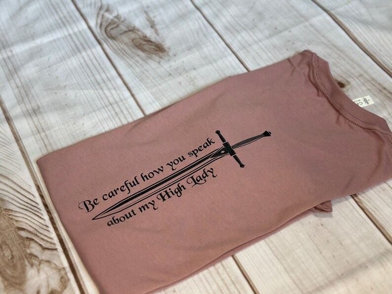 May include: A mauve t-shirt with a black graphic of a sword and the text "Be careful how you speak about my High Lady".