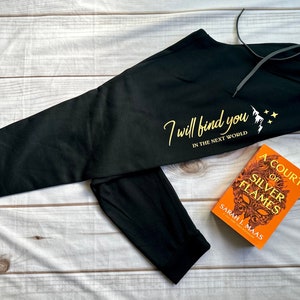 K&ouml;nnte beinhalten: Schwarze Jogginghose mit Kordelzug und goldenem Text "I will find you in the next world". Neben der Hose liegt ein Exemplar von "A Court of Silver Flames" von Sarah J. Maas.