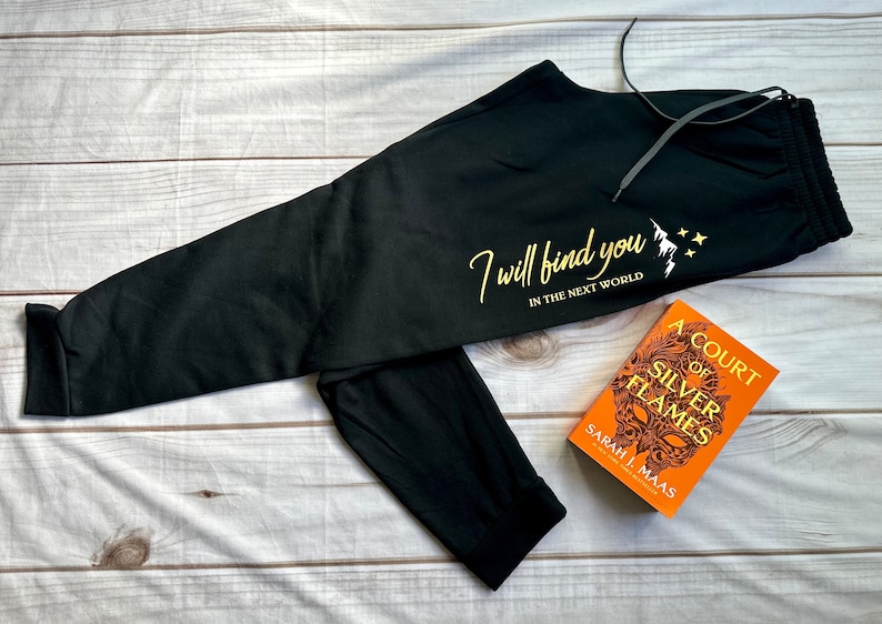 K&ouml;nnte beinhalten: Schwarze Jogginghose mit Kordelzug und goldenem Text "I will find you in the next world". Neben der Hose liegt ein Exemplar von "A Court of Silver Flames" von Sarah J. Maas.