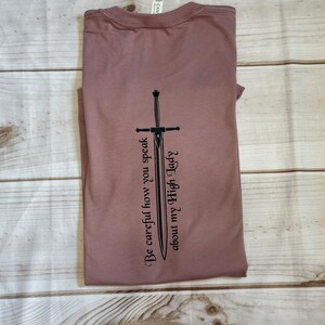 May include: A mauve t-shirt with a black sword graphic and the text "Be careful how you speak about my High Lady".
