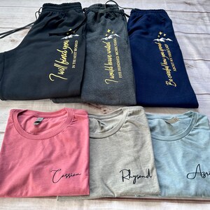 K&ouml;nnte beinhalten: Eine Auswahl an gefalteter Kleidung, darunter Jogginghosen und T-Shirts. Die Jogginghosen sind marineblau, anthrazit und schwarz, jeweils mit goldenem Text. Die T-Shirts sind staubrosa, grau und hellblau, jeweils mit einem Namen in schwarzer Schrift.