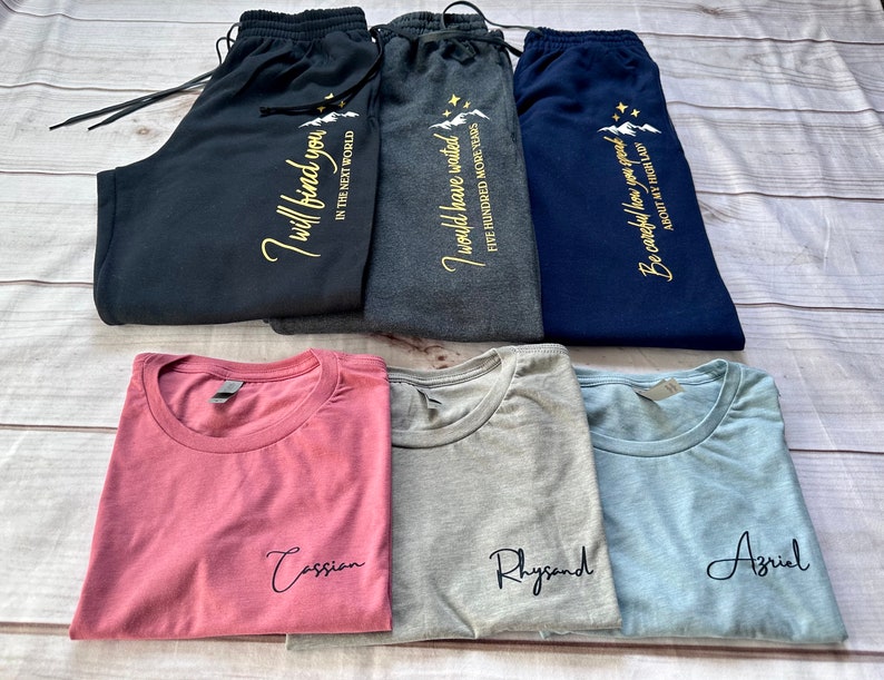 K&ouml;nnte beinhalten: Eine Auswahl an gefalteter Kleidung, darunter Jogginghosen und T-Shirts. Die Jogginghosen sind marineblau, anthrazit und schwarz, jeweils mit goldenem Text. Die T-Shirts sind staubrosa, grau und hellblau, jeweils mit einem Namen in schwarzer Schrift.