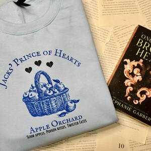 May include: A light blue sweatshirt with a blue graphic print of a basket of apples and the text "Jack's? Prince of Hearts Apple Orchard Sour Apples, Poison Kisses, Twisted Fates". The sweatshirt is lying on a surface with a book titled "Once Upon a Broken Heart" by Stephanie Garber.