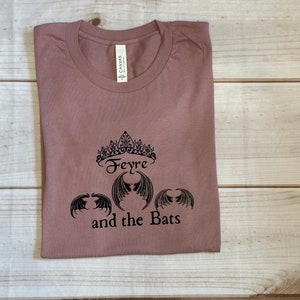 May include: A mauve t-shirt with a black graphic design featuring a crown, the word "Feyre", and three bat wings. The text "and the Bats" is below the graphic.