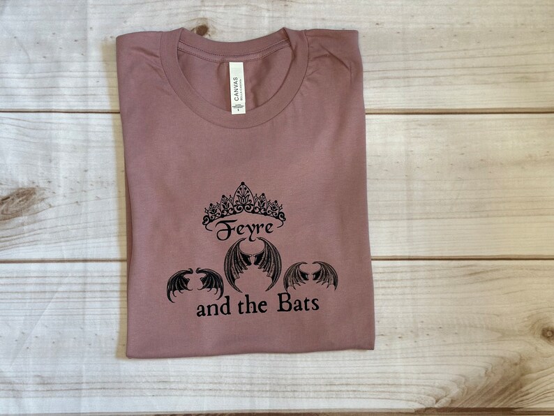 May include: A mauve t-shirt with a black graphic design featuring a crown, the word "Feyre", and three bat wings. The text "and the Bats" is below the graphic.