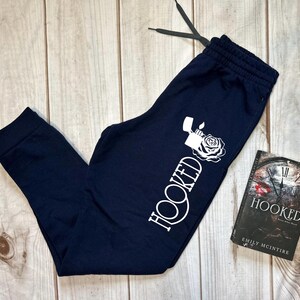 May include: Navy blue sweatpants with a white graphic of a lighter and rose, and the word "HOOKED". A book titled "HOOKED" by Emily McIntire is next to the sweatpants. The sweatpants have a drawstring.