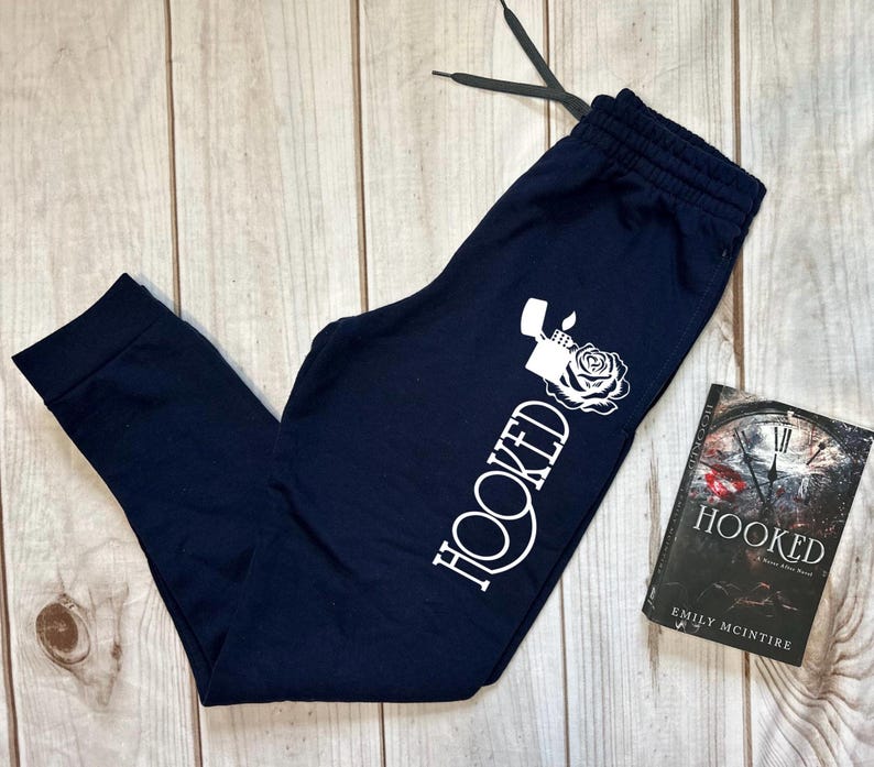 May include: Navy blue sweatpants with a white graphic of a lighter and rose, and the word "HOOKED". A book titled "HOOKED" by Emily McIntire is next to the sweatpants. The sweatpants have a drawstring.