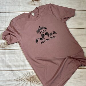 May include: A mauve t-shirt with a black graphic print of a crown, the word "Feyre", and bats with the text "and the Bats".
