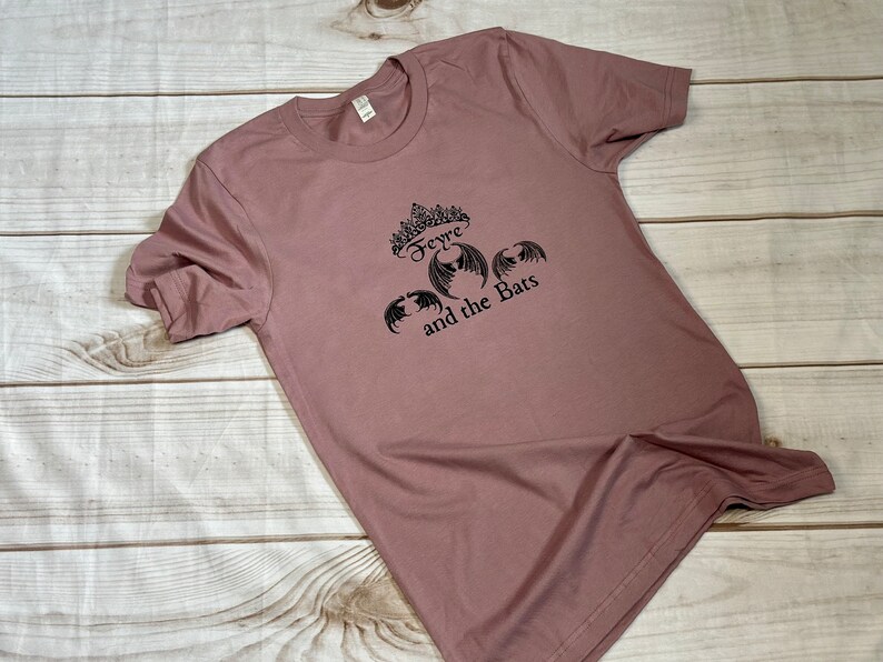 May include: A mauve t-shirt with a black graphic print of a crown, the word "Feyre", and bats with the text "and the Bats".