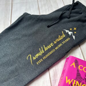 K&ouml;nnte beinhalten: Dunkelgraue Jogginghose mit Kordelzug und goldenem Text "I would have waited FIVE HUNDRED MORE YEARS" mit Berg- und Sternmotiv. Ein rosa Buch mit wei&szlig;em Text ist sichtbar.
