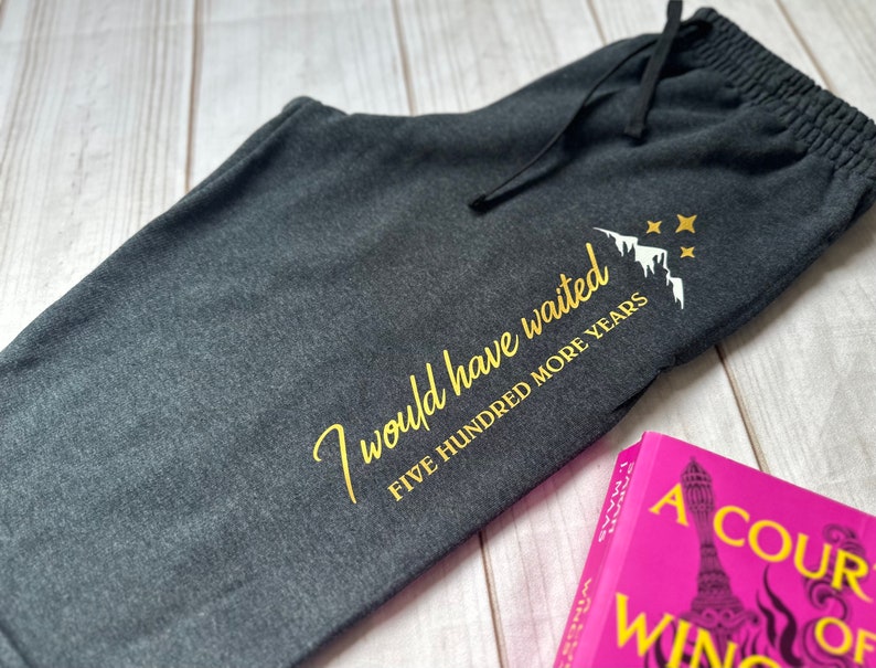 K&ouml;nnte beinhalten: Dunkelgraue Jogginghose mit Kordelzug und goldenem Text "I would have waited FIVE HUNDRED MORE YEARS" mit Berg- und Sternmotiv. Ein rosa Buch mit wei&szlig;em Text ist sichtbar.