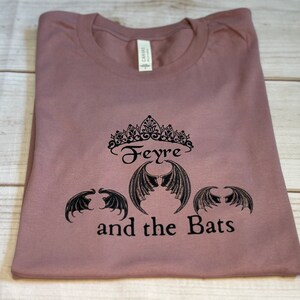 May include: A mauve t-shirt with a black graphic design featuring a crown, the word "Feyre", and three bat wings. The text "and the Bats" is below the wings.