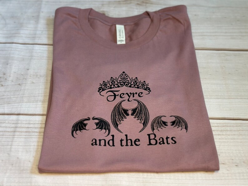 May include: A mauve t-shirt with a black graphic design featuring a crown, the word "Feyre", and three bat wings. The text "and the Bats" is below the wings.