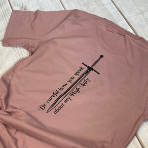 May include: A dusty rose t-shirt with a black graphic of a sword and the text "Be careful how you speak about my High Lady".