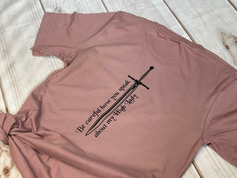 May include: A dusty rose t-shirt with a black graphic of a sword and the text "Be careful how you speak about my High Lady".