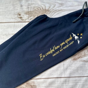 K&ouml;nnte beinhalten: Marineblaue Jogginghose mit Kordelzug. Goldener Text: "Be careful how you speak ABOUT MY HIGH LADY" mit Berg- und Stern-Grafik. Die Hose ist aus weichem Material gefertigt.
