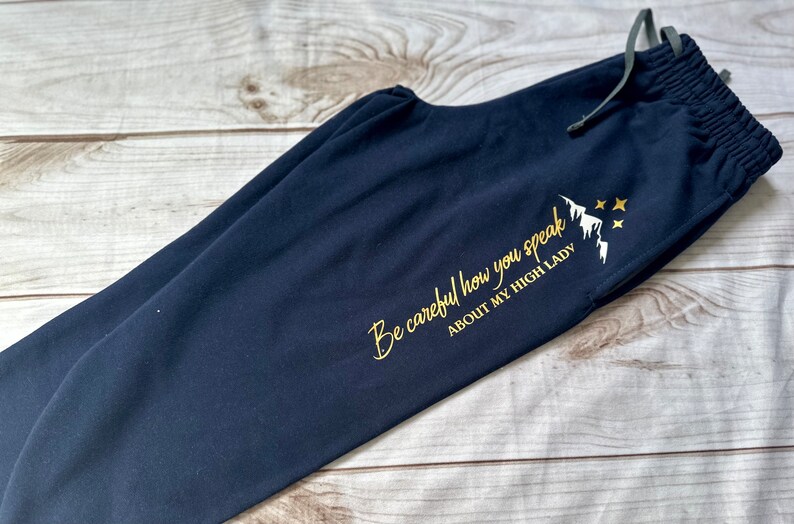 K&ouml;nnte beinhalten: Marineblaue Jogginghose mit Kordelzug. Goldener Text: "Be careful how you speak ABOUT MY HIGH LADY" mit Berg- und Stern-Grafik. Die Hose ist aus weichem Material gefertigt.