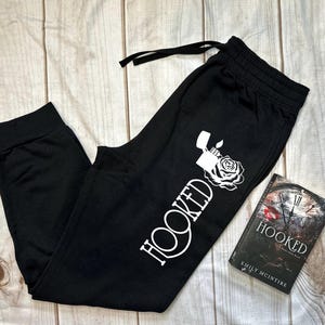 May include: Black sweatpants with a white graphic that says "HOOKED" with a lighter and rose design. A book titled "HOOKED" by Emily McIntire is next to the sweatpants. The sweatpants have a drawstring and elastic waistband.