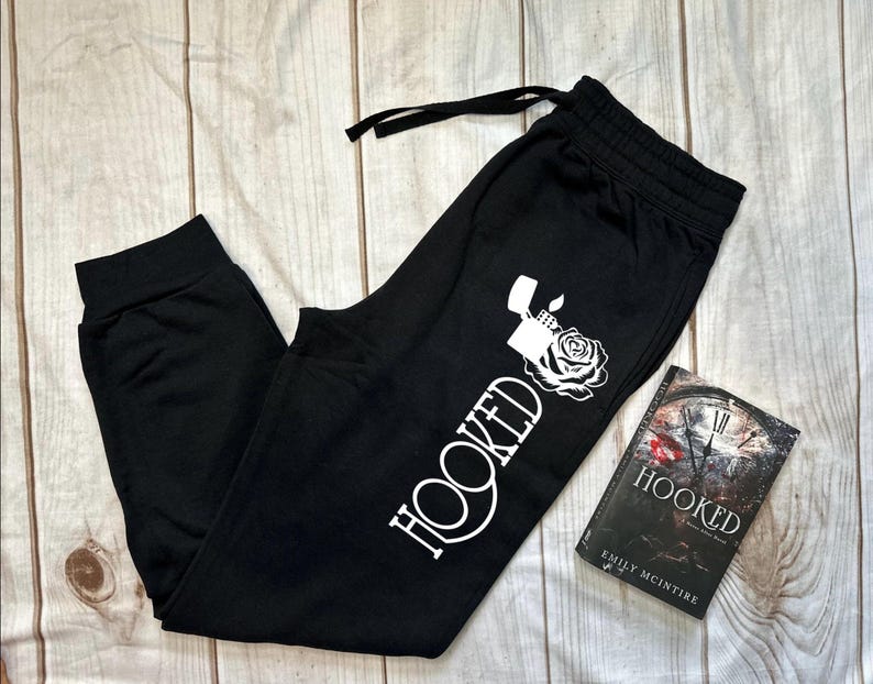 May include: Black sweatpants with a white graphic that says "HOOKED" with a lighter and rose design. A book titled "HOOKED" by Emily McIntire is next to the sweatpants. The sweatpants have a drawstring and elastic waistband.