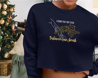 Puede incluir: Sudadera corta azul marino con texto dorado y un gráfico de manos sosteniendo sobres. El texto dice "A bond that not even distance can break". Un árbol de Navidad es visible en el fondo.