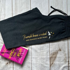 K&ouml;nnte beinhalten: Dunkelgraue Jogginghose mit Kordelzug und goldenem Text "I would have waited FIVE HUNDRED MORE YEARS" mit einem Berg- und Stern-Motiv. Ein Buch mit pinkfarbenem Einband ist ebenfalls im Bild.