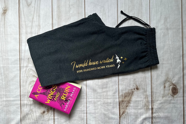 K&ouml;nnte beinhalten: Dunkelgraue Jogginghose mit Kordelzug und goldenem Text "I would have waited FIVE HUNDRED MORE YEARS" mit einem Berg- und Stern-Motiv. Ein Buch mit pinkfarbenem Einband ist ebenfalls im Bild.