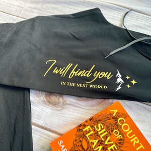 K&ouml;nnte beinhalten: Schwarze Jogginghose mit Kordelzug und dem Text "I will find you in the next world" in goldener Schrift. Unter dem Text befindet sich eine wei&szlig;e Berg- und Sternengrafik. Ein Buch mit orangefarbenem Einband ist im Vordergrund.