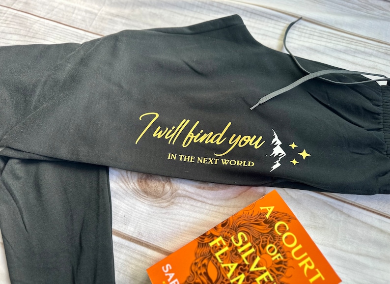 K&ouml;nnte beinhalten: Schwarze Jogginghose mit Kordelzug und dem Text "I will find you in the next world" in goldener Schrift. Unter dem Text befindet sich eine wei&szlig;e Berg- und Sternengrafik. Ein Buch mit orangefarbenem Einband ist im Vordergrund.