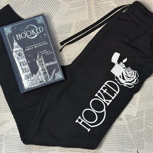 May include: Black sweatpants with a white graphic of a lighter and rose, and the word "HOOKED". A hardcover book titled "HOOKED" by Emily McIntire is also in the image. The book cover features a London cityscape.
