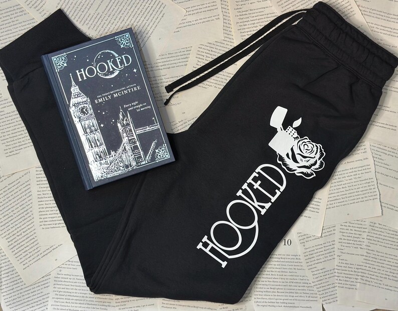 May include: Black sweatpants with a white graphic of a lighter and rose, and the word "HOOKED". A hardcover book titled "HOOKED" by Emily McIntire is also in the image. The book cover features a London cityscape.