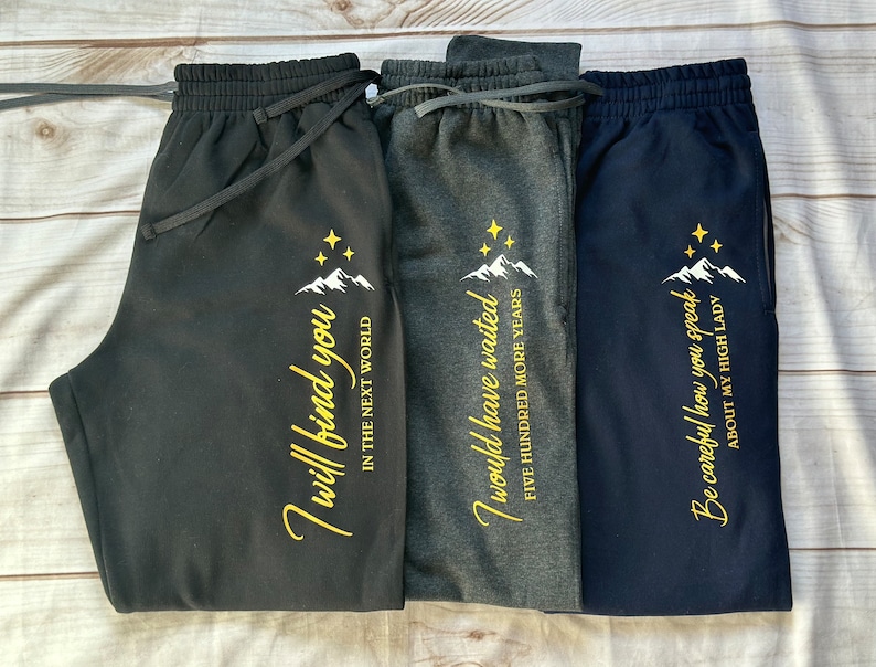 K&ouml;nnte beinhalten: Drei Sweatpants in Schwarz, Grau und Marineblau. Jede Hose hat eine Berg- und Sternengrafik mit goldenem Text. Der Text auf jeder Hose lautet: "I will find you", "I would have waited" und "Be careful how you speak."