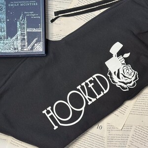 May include: Black sweatpants with a white graphic of a lighter and rose, with the word "HOOKED" in white. A book with a blue cover and the title "Every night and straight on 'til morning" is on top of the sweatpants.
