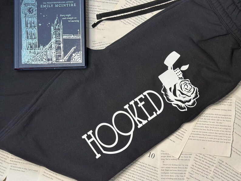 May include: Black sweatpants with a white graphic of a lighter and rose, with the word "HOOKED" in white. A book with a blue cover and the title "Every night and straight on 'til morning" is on top of the sweatpants.