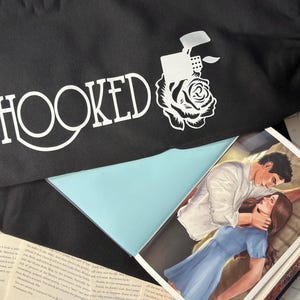 May include: Black fabric with the word "HOOKED" in white, along with a graphic of a lighter and rose. A book with a couple in a romantic embrace is visible, with pages of text surrounding it.