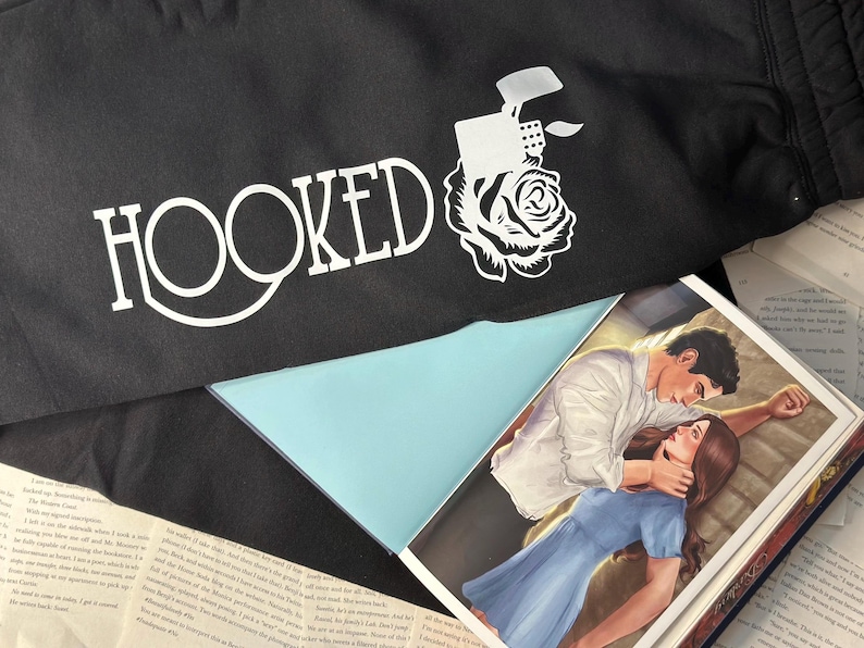 May include: Black fabric with the word "HOOKED" in white, along with a graphic of a lighter and rose. A book with a couple in a romantic embrace is visible, with pages of text surrounding it.