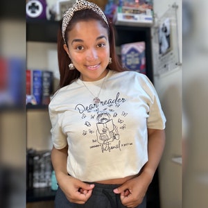 May include: A beige t-shirt with a black graphic of a person reading a book with butterflies flying around them. The text on the shirt reads "Dear reader, pick somewhere fictional & just run."