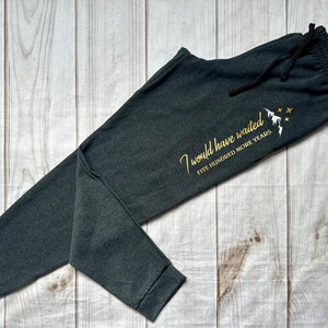 K&ouml;nnte beinhalten: Dunkelgraue Jogginghose mit Kordelzug. Der Text "I would have waited FIVE HUNDRED MORE YEARS" ist in Gold mit einem Berg- und Sternmotiv aufgedruckt. Die Hose liegt auf einer hellen Holzmaserung.