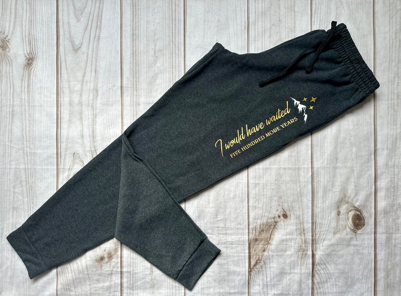 K&ouml;nnte beinhalten: Dunkelgraue Jogginghose mit Kordelzug. Der Text "I would have waited FIVE HUNDRED MORE YEARS" ist in Gold mit einem Berg- und Sternmotiv aufgedruckt. Die Hose liegt auf einer hellen Holzmaserung.