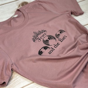 May include: A mauve t-shirt with a black graphic print of a crown with the text "Feyre and the Bats".