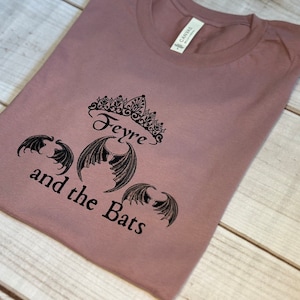 May include: A mauve t-shirt with a black graphic design featuring a crown, the word "Feyre", and three bat wings. The text "and the Bats" is written below the graphic.