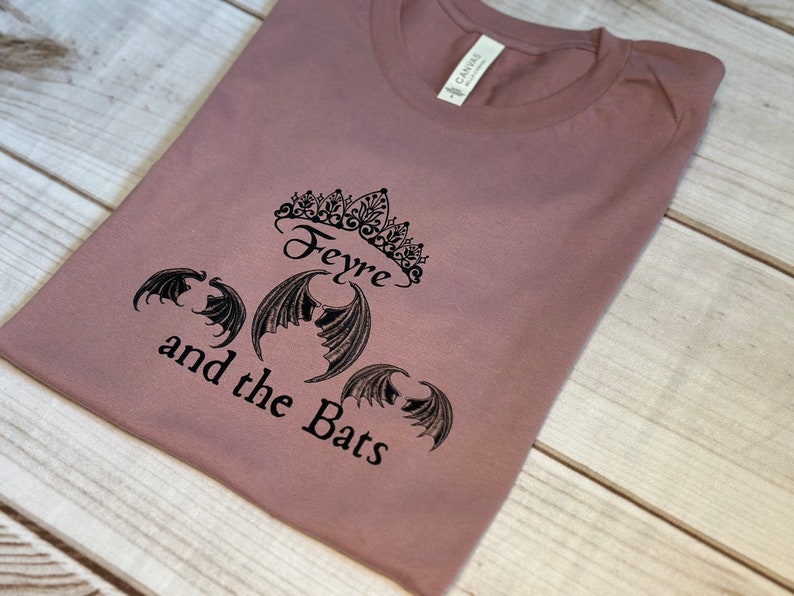 May include: A mauve t-shirt with a black graphic design featuring a crown, the word "Feyre", and three bat wings. The text "and the Bats" is written below the graphic.