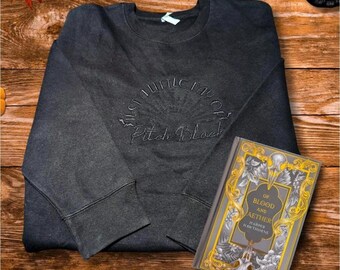 Puede incluir: Sudadera gris oscuro con el texto bordado "Just a Little Fau of Pitch Black". Junto a la sudadera hay un libro titulado "Of Blood and Aether" de Harper Hawthorne. El libro tiene una cubierta dorada y gris.